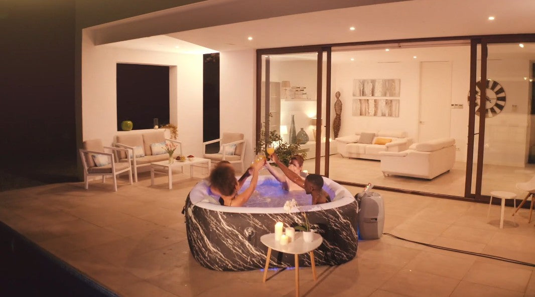 People in a hot tub on a patio at night with a modern house interior visible.