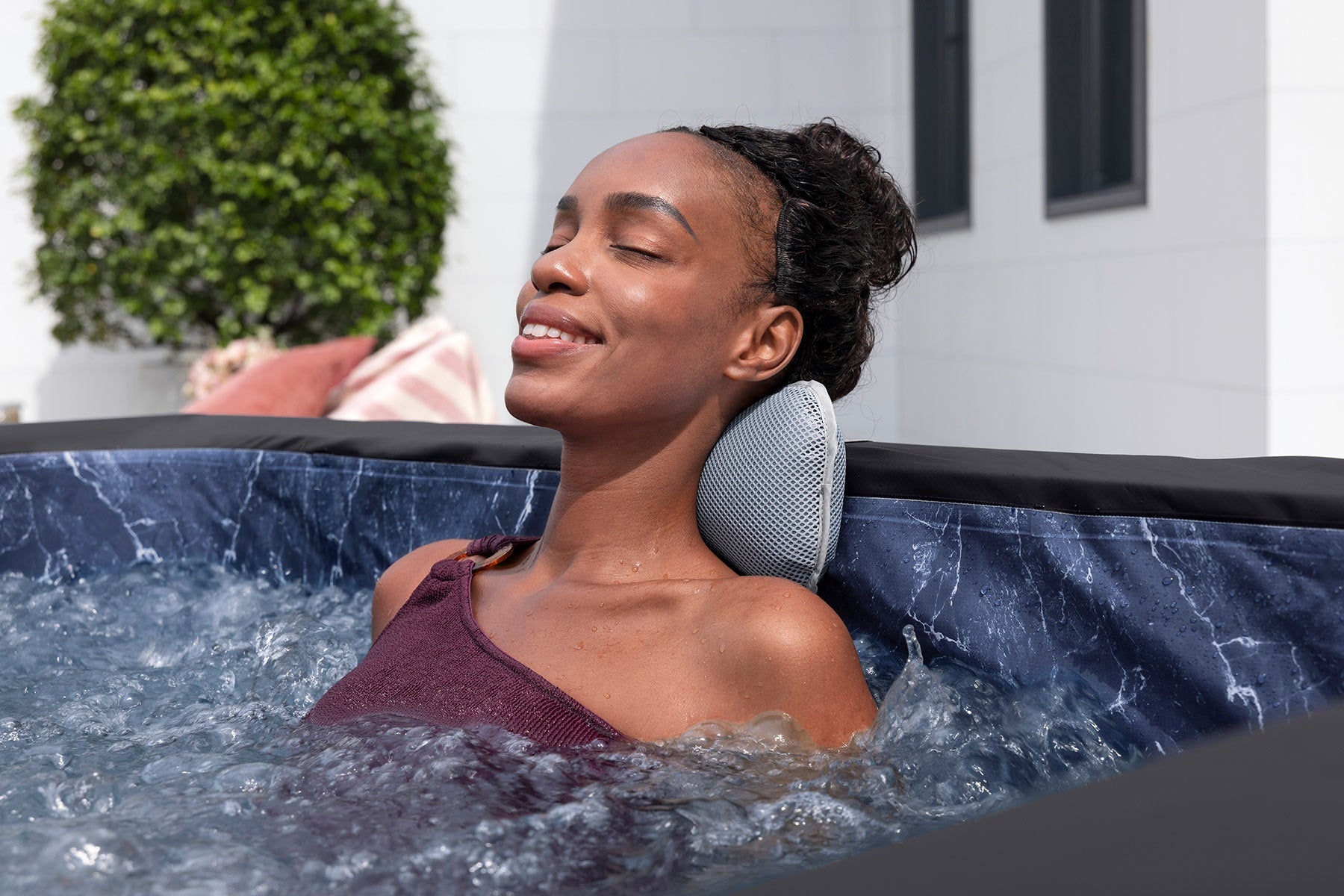 Woman relaxing in a hot tub with a neck pillow, surrounded by greenery and a building.
