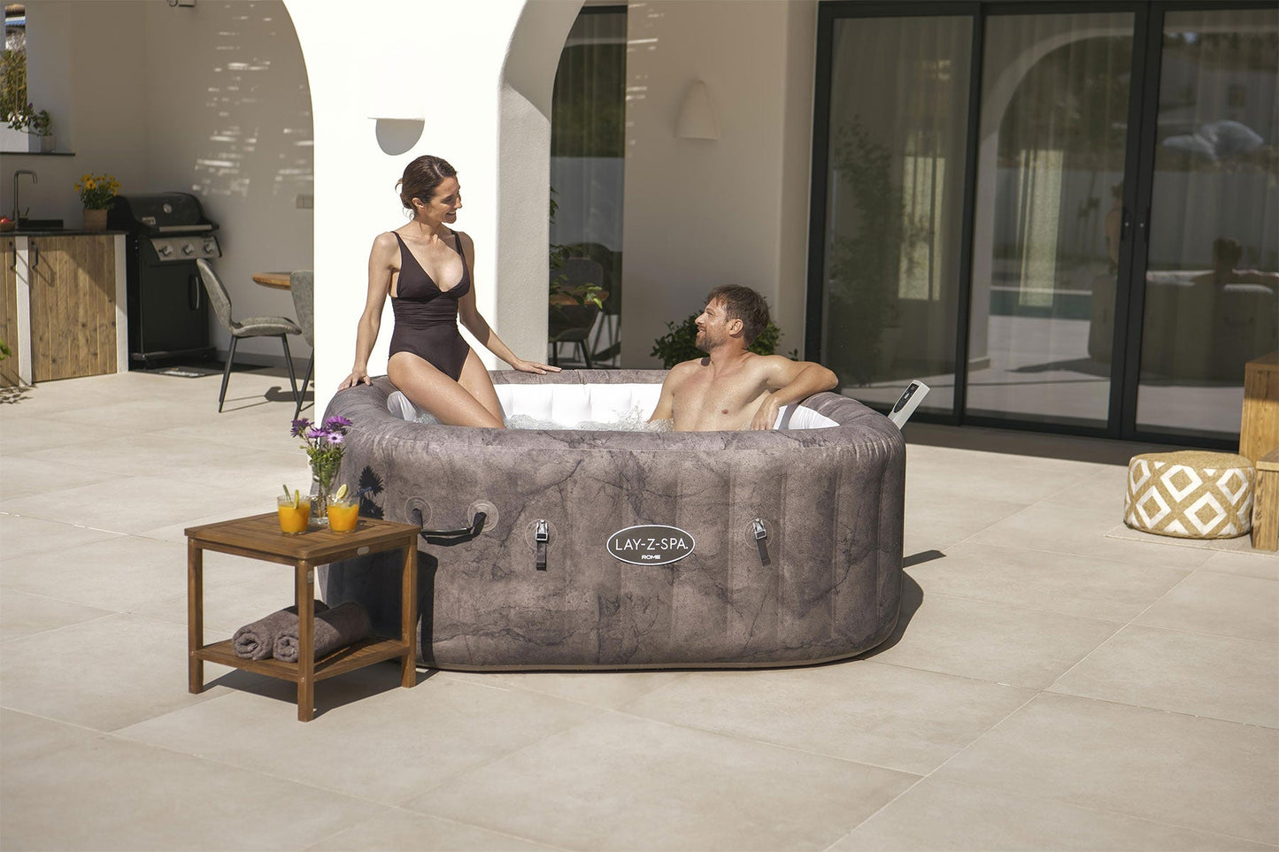 Two people in a Lay-Z-Spa hot tub on a patio with a table and chairs nearby.