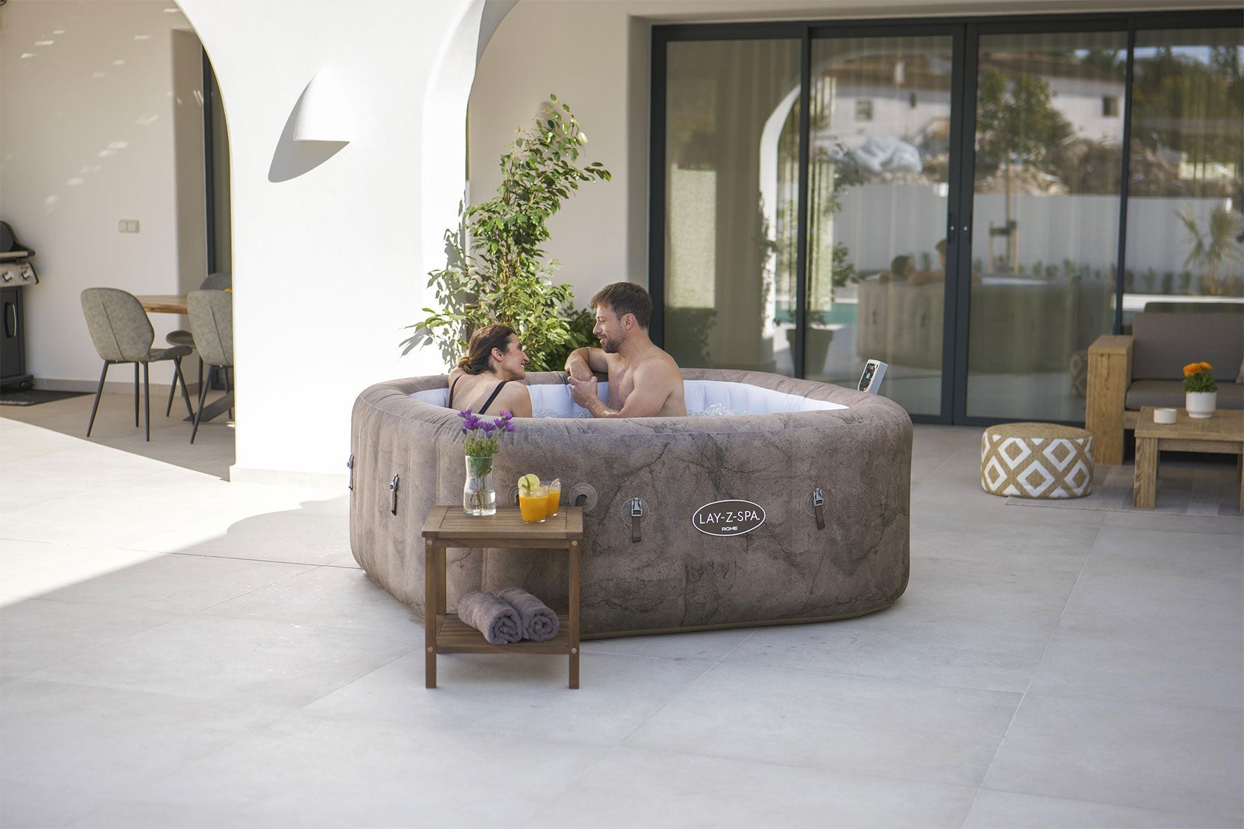 Two people in a Lay-Z-Spa hot tub on a patio with a table and chairs nearby.