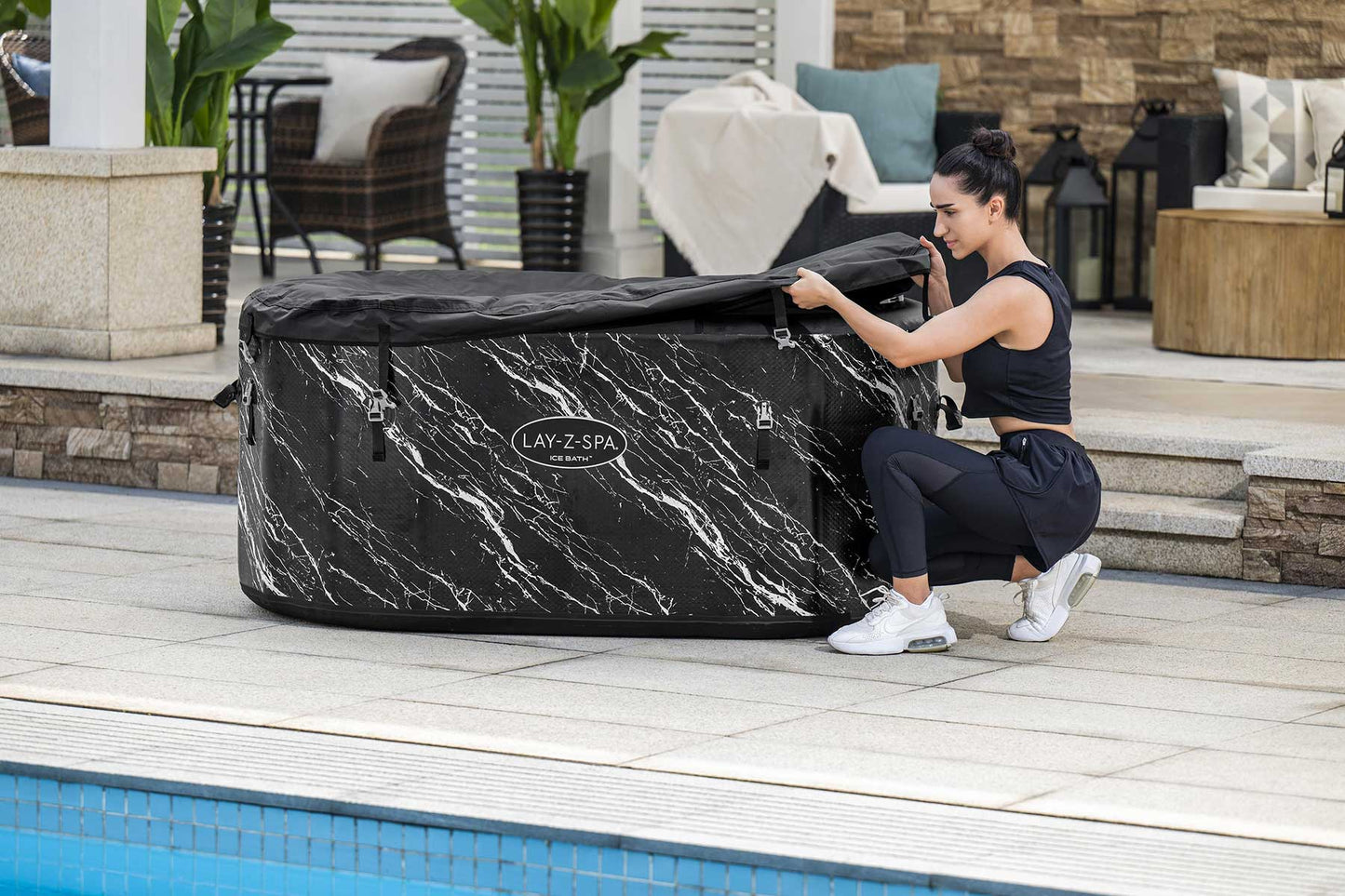 Woman opening a black marble-patterned hot tub on a patio by a pool.