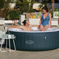 Two people in an inflatable hot tub with a 'Lay-Z-Spa' brand logo, surrounded by outdoor furniture and plants.