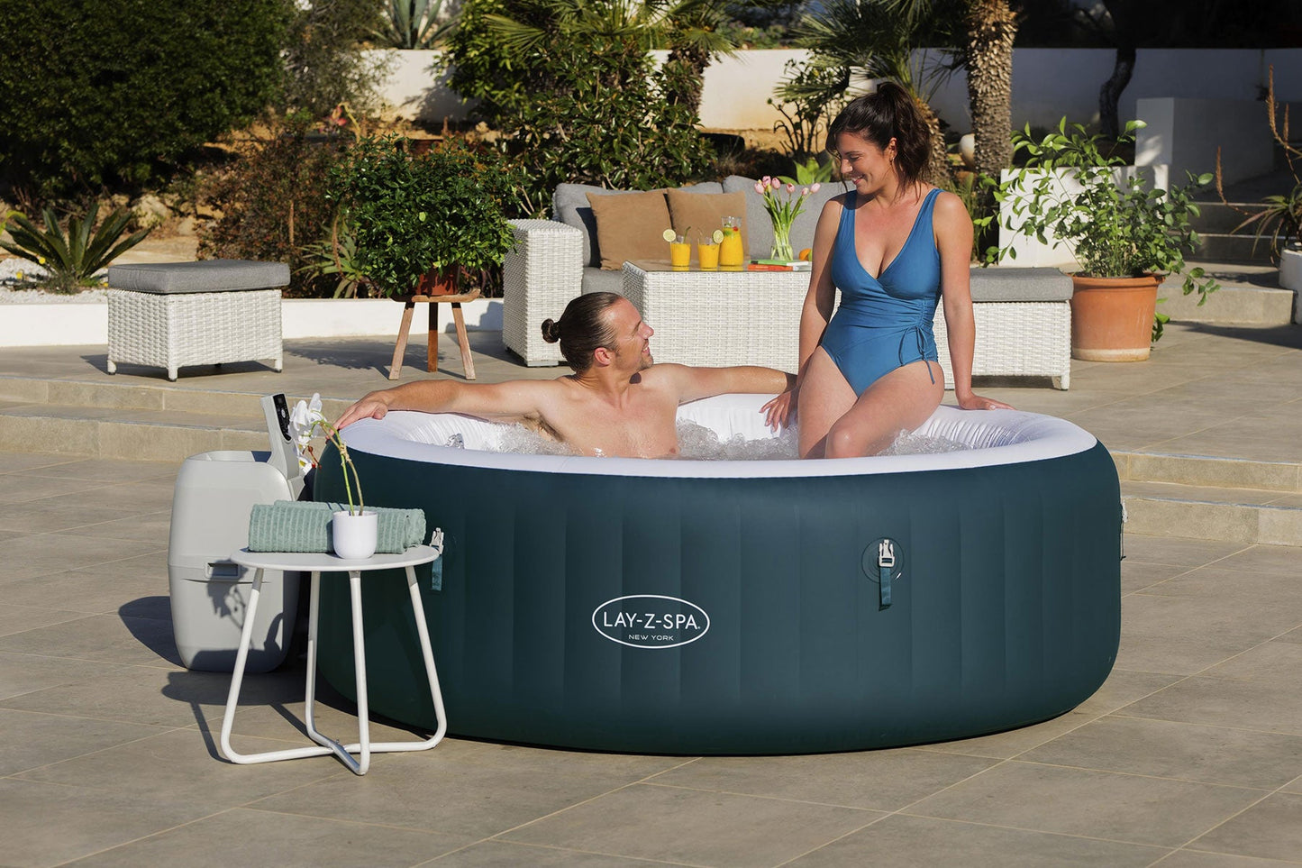 Two people in an inflatable hot tub with a 'Lay-Z-Spa' brand logo, surrounded by outdoor furniture and plants.