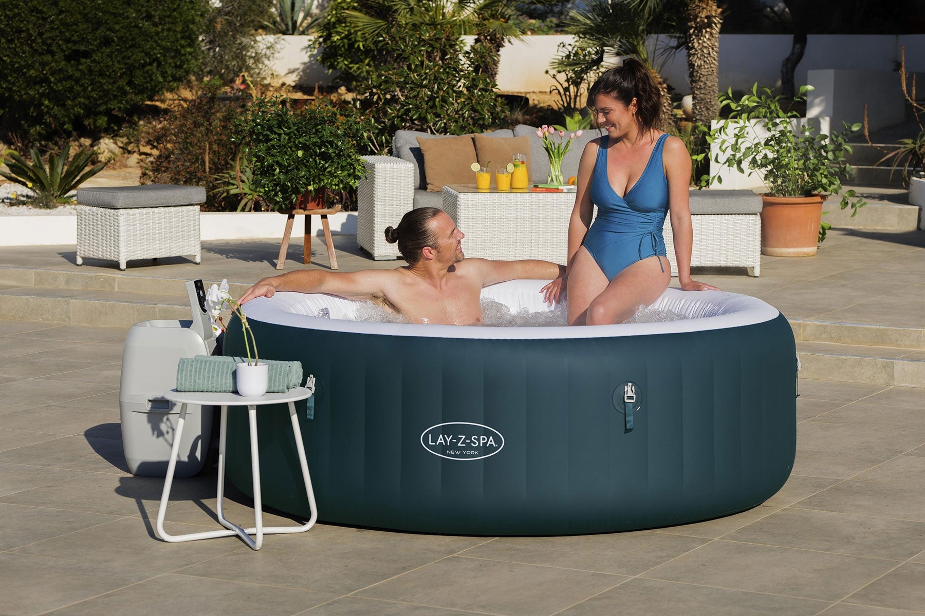 Two people in an inflatable hot tub with a 'Lay-Z-Spa' brand logo, surrounded by outdoor furniture and plants.