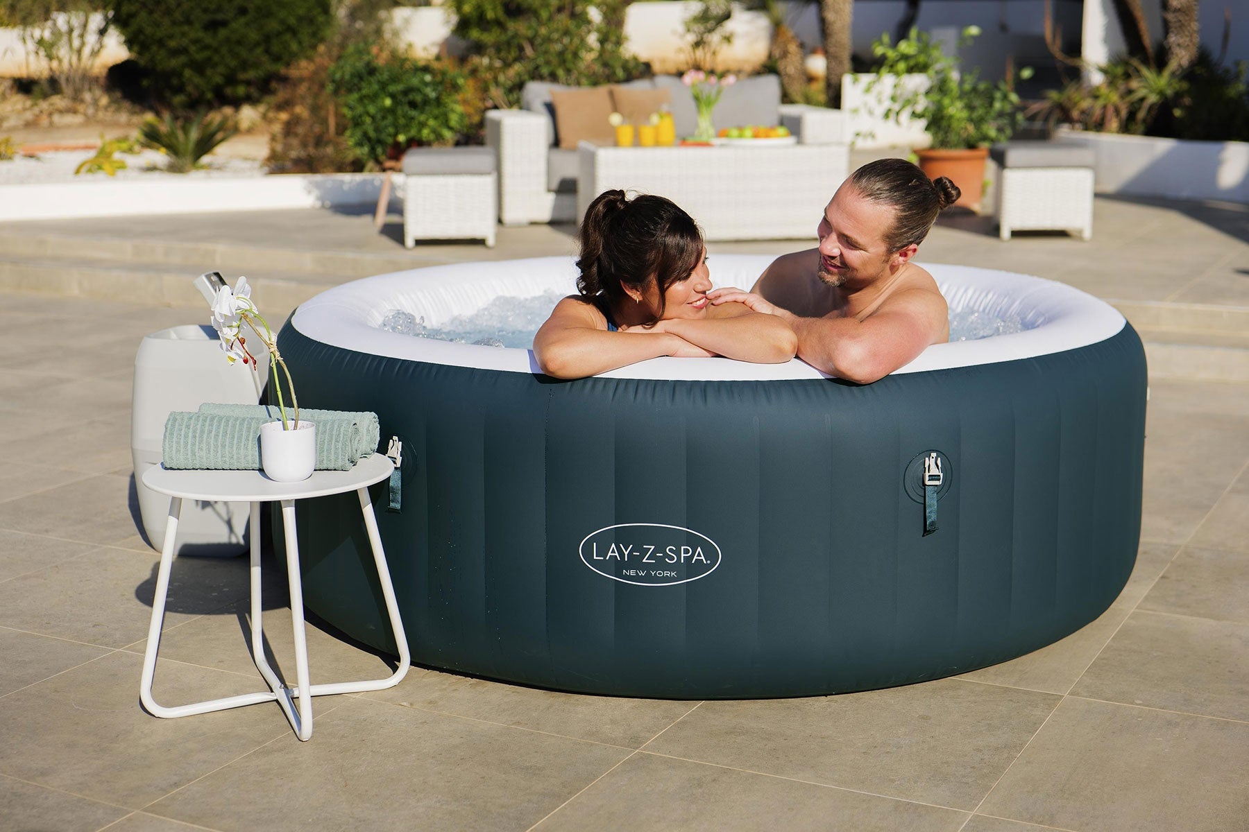 Two people in a Lay-Z-Spa inflatable hot tub on a patio with outdoor furniture.
